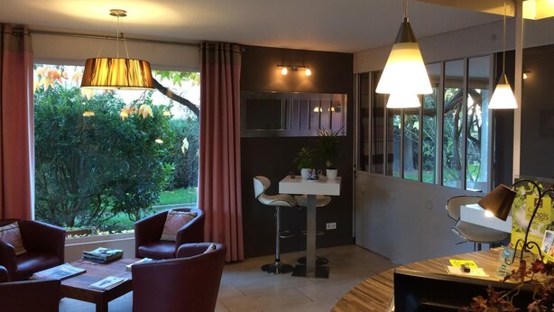 Ett modernt vardagsrum med en soffgrupp, ett soffbord och ett skrivbord med en lampa. Det finns ett fönster med gardiner, en växt och en utsikt över grönområden utanför.
