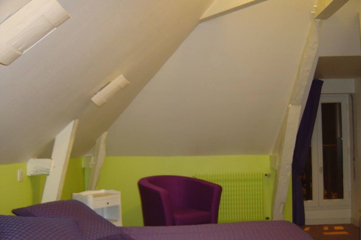 A room with a sloped ceiling, a bed with a purple bedspread, a purple chair, and a white bedside table.