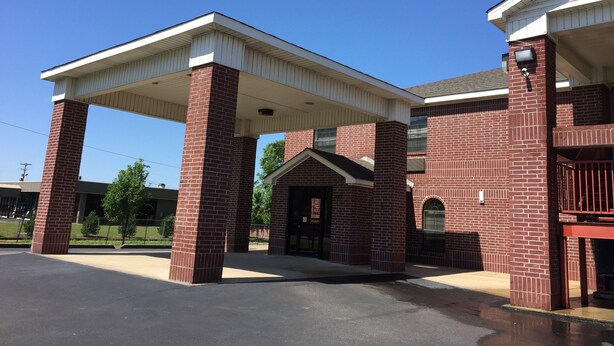 A building with a covered entrance area supported by brick pillars, a clear sky, and a parking lot. - Sunset Inn (West Memphis)