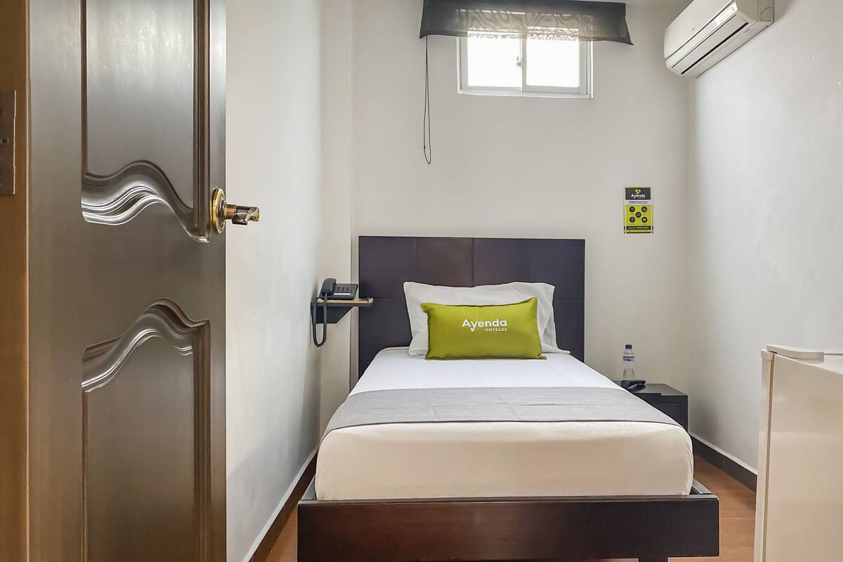 A hotel room with a bed, a bedside table with a phone, a water bottle, and a sign indicating the hotel name.