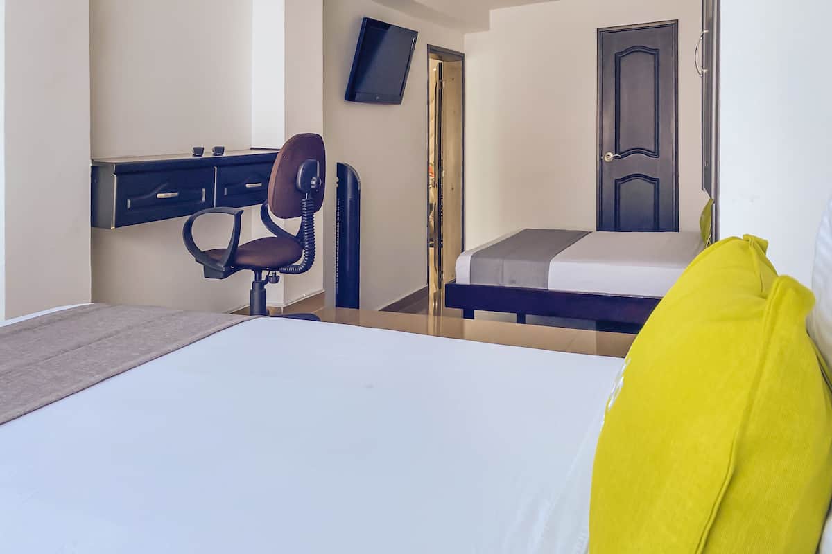A bedroom with a bed, a desk with a chair, a TV mounted on the wall, and a door.