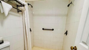 Um banheiro pequeno e branco, com vaso sanitário, chuveiro com porta de vidro e piso de azulejos brancos.