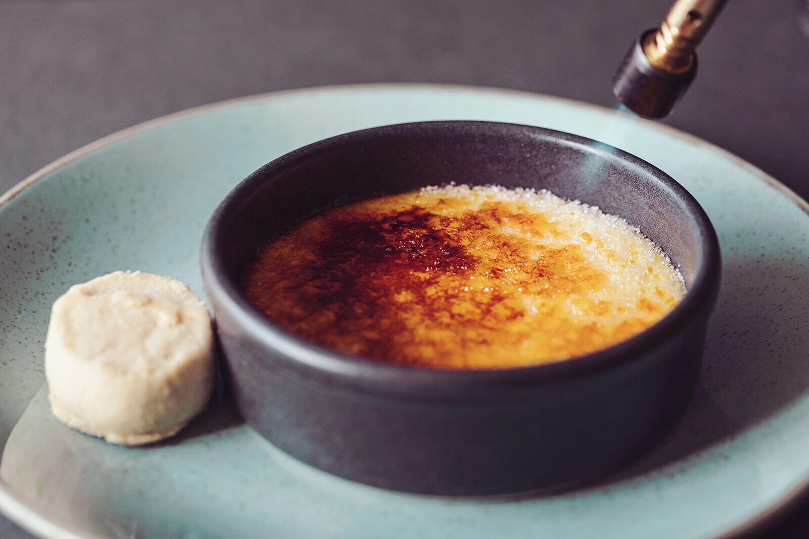 Un bol de crema brûlée sobre un plato con una capa caramelizada, acompañado de una pequeña cucharada de crema.