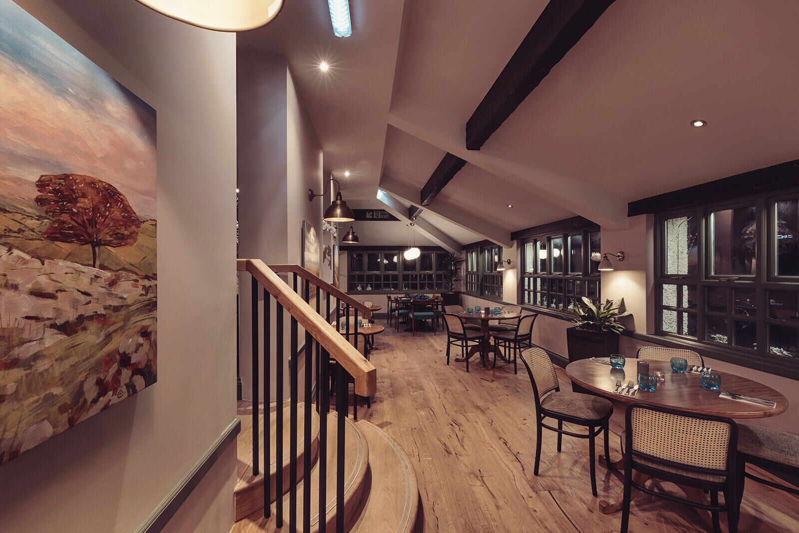 A spacious dining area with wooden flooring, a staircase with black railings, and a large window providing natural light.