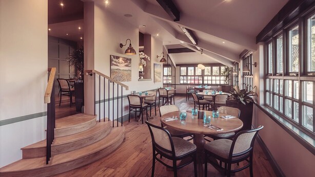 Un comedor amplio con mesas y sillas de madera, una escalera que sube a un nivel superior y ventanas grandes que dejan entrar luz natural.