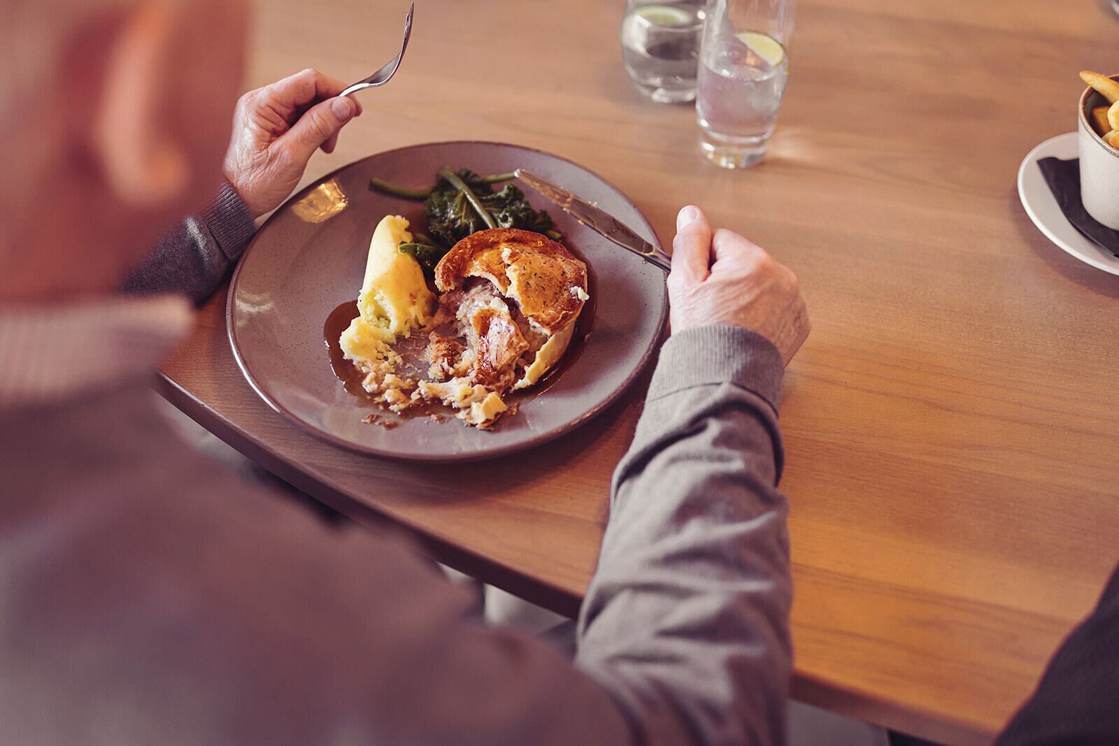 Una persona cenando en una mesa de madera, con un plato de comida, un tenedor y un vaso de agua.