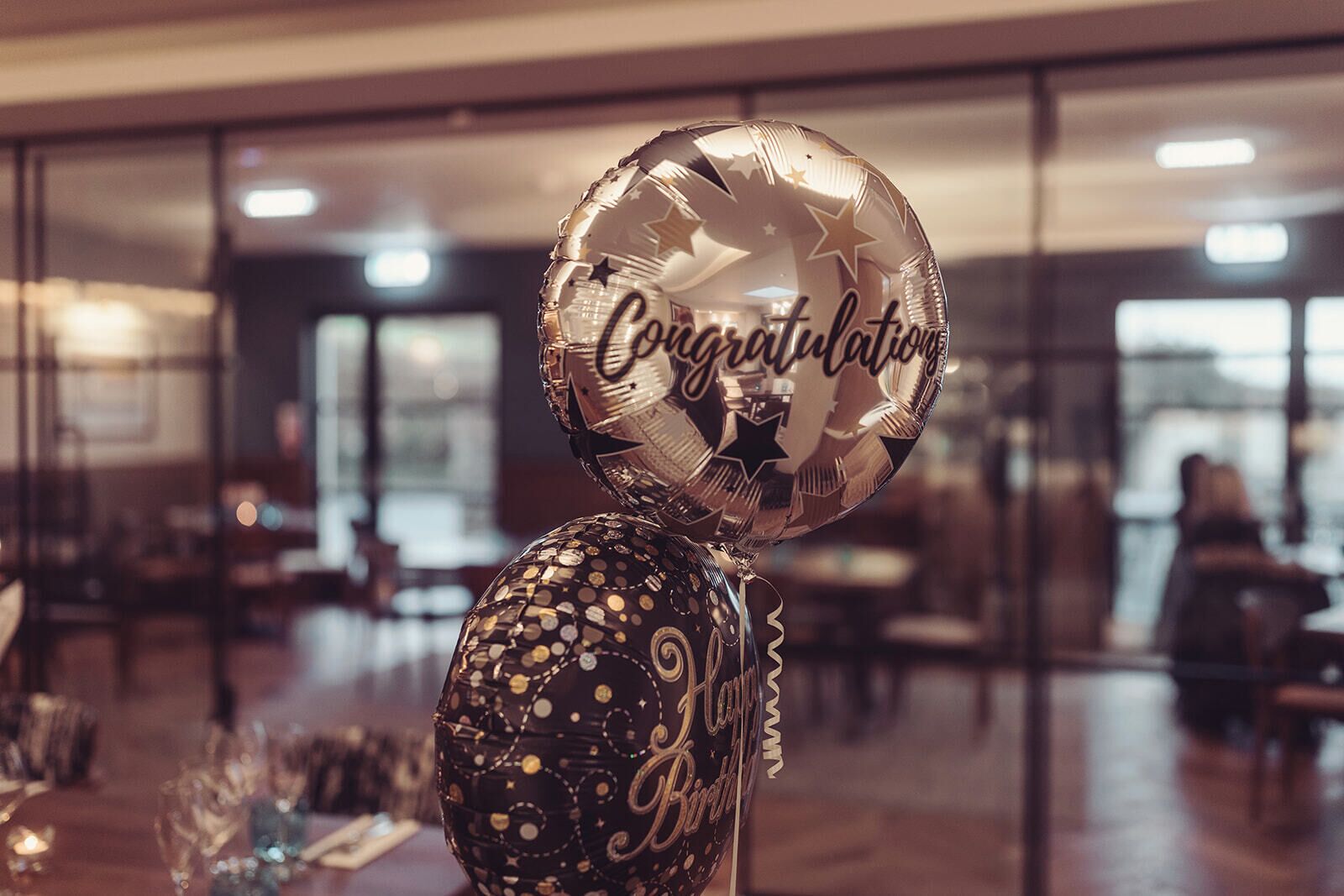 Una habitación con un separador de vidrio, una mesa con un vaso y un globo que dice 'Felicidades'.