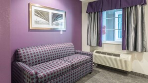 Una habitación con pared morada, un sofá estampado, un cuadro enmarcado, una ventana con cortinas y un aire acondicionado instalado en la pared.