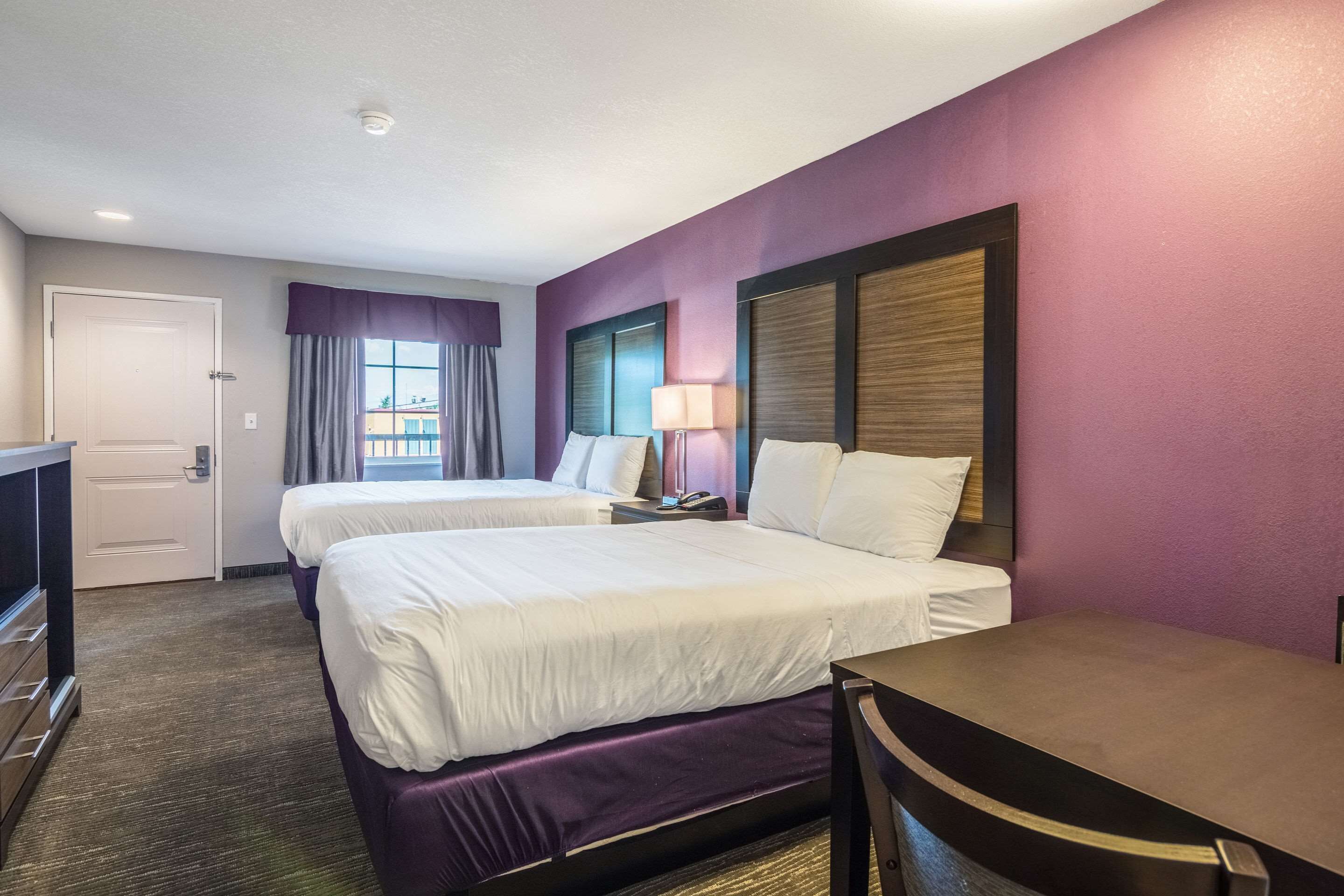 A hotel room with two beds, a desk, a TV, and a window with curtains.