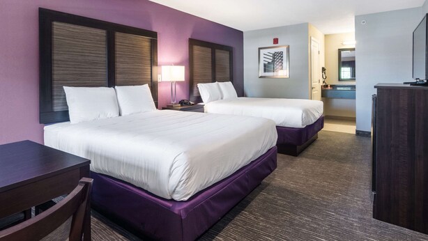 Habitación de hotel con dos camas, un escritorio, una silla, una lámpara, un baño con espejo y un televisor.