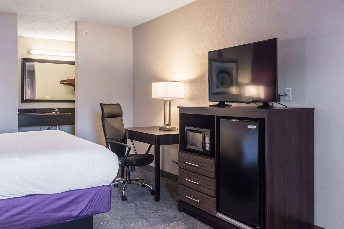 A hotel room with a bed, a desk with a lamp, a television, and a minibar.