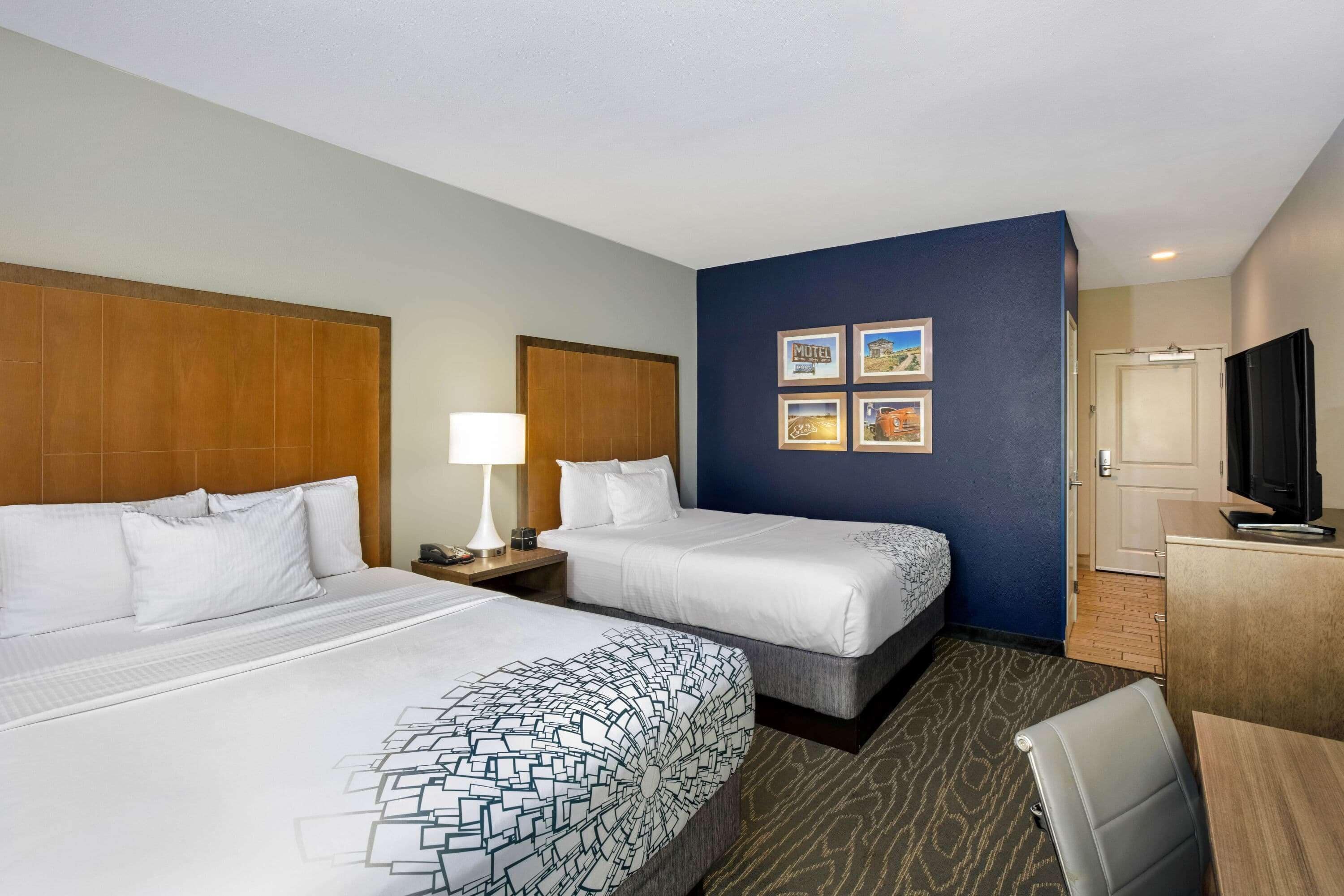A hotel room with two beds, a desk, a chair, a TV, and a wall with framed pictures.