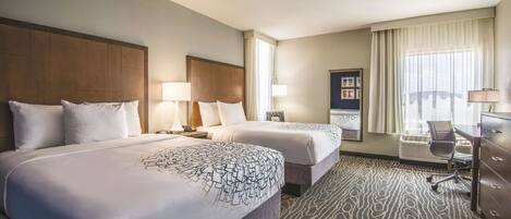 A hotel room with two beds, a desk, a chair, and a large window with curtains.