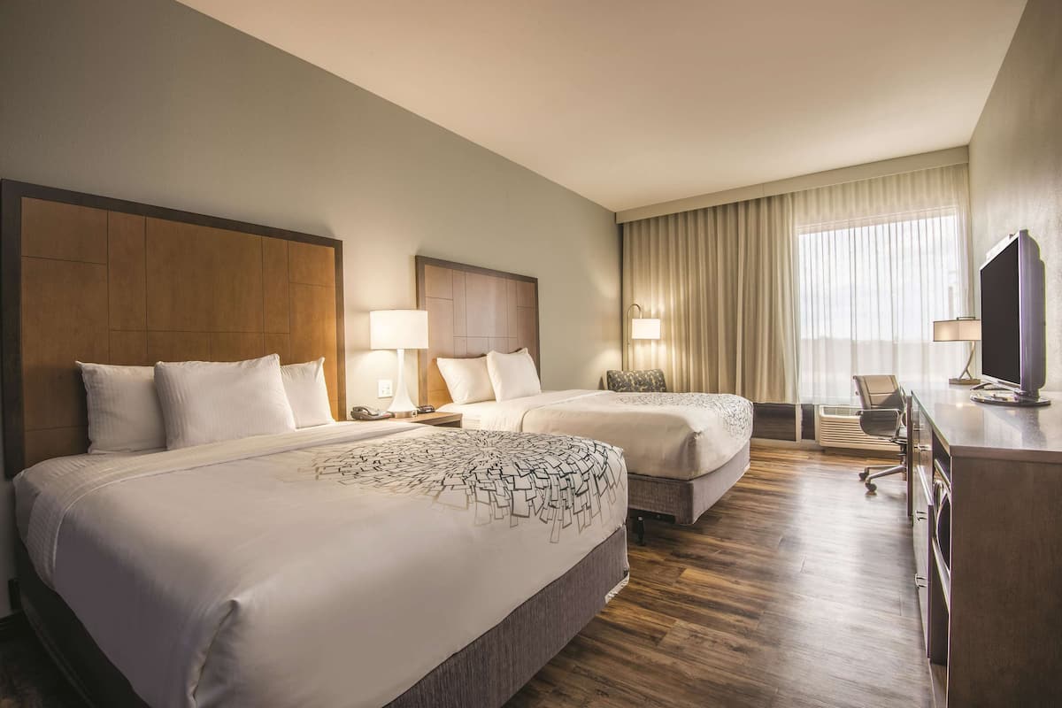 Habitación de hotel con dos camas, un escritorio con televisor y un ventanal con cortinas.