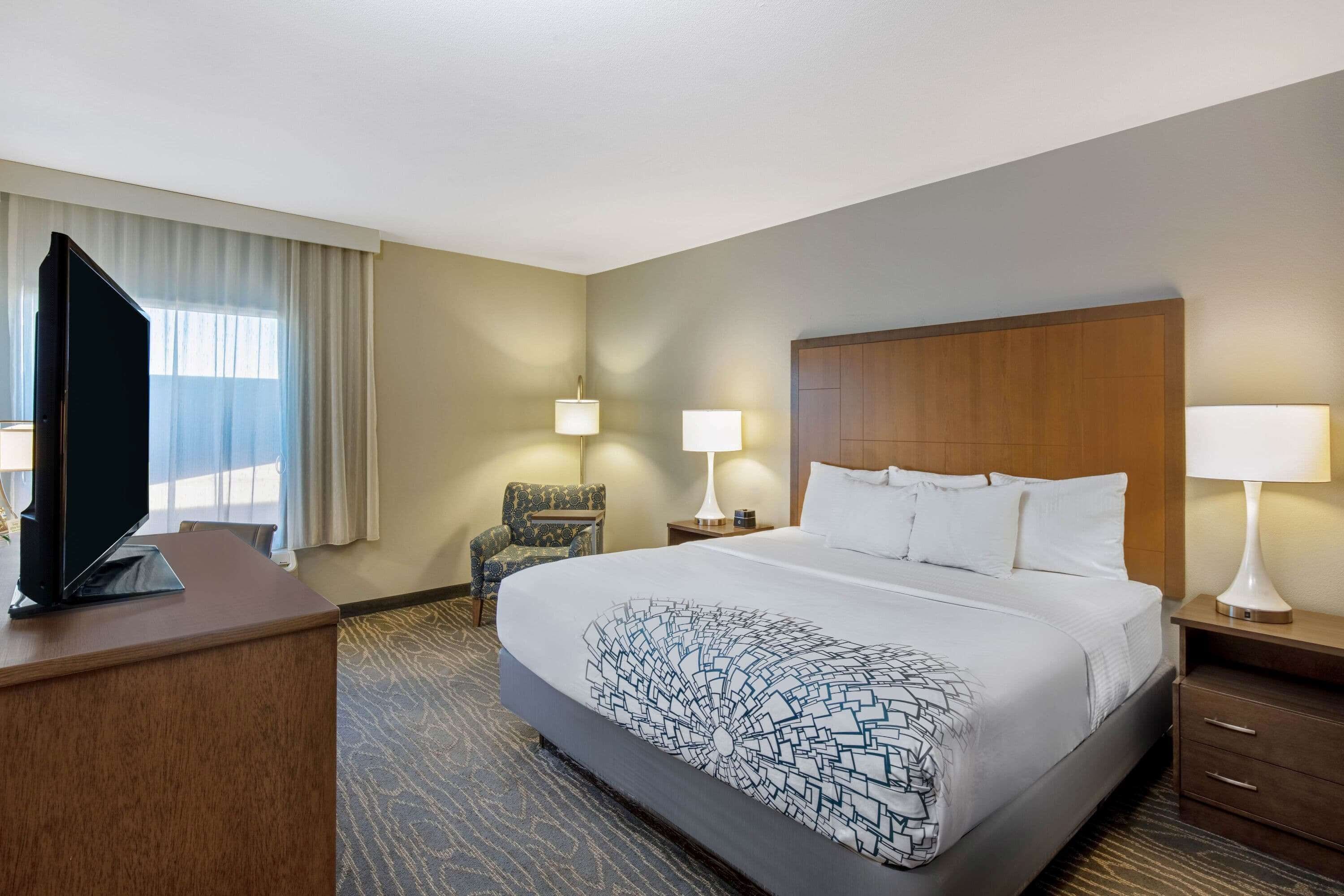 Habitación de hotel con una cama grande, un televisor sobre un escritorio, dos lámparas de noche, una silla y una ventana con cortinas.
