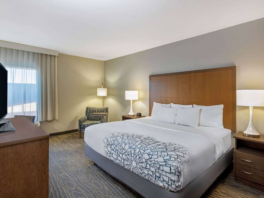 A hotel room with a large bed, a television on a desk, two bedside lamps, a chair, and a window with curtains.