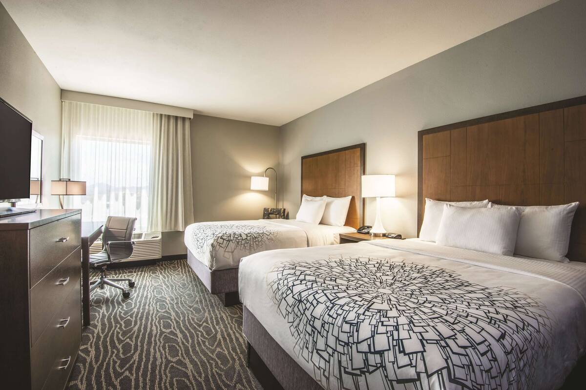 Habitación de hotel con dos camas, un escritorio, una silla, un aparador y un ventanal con cortinas.