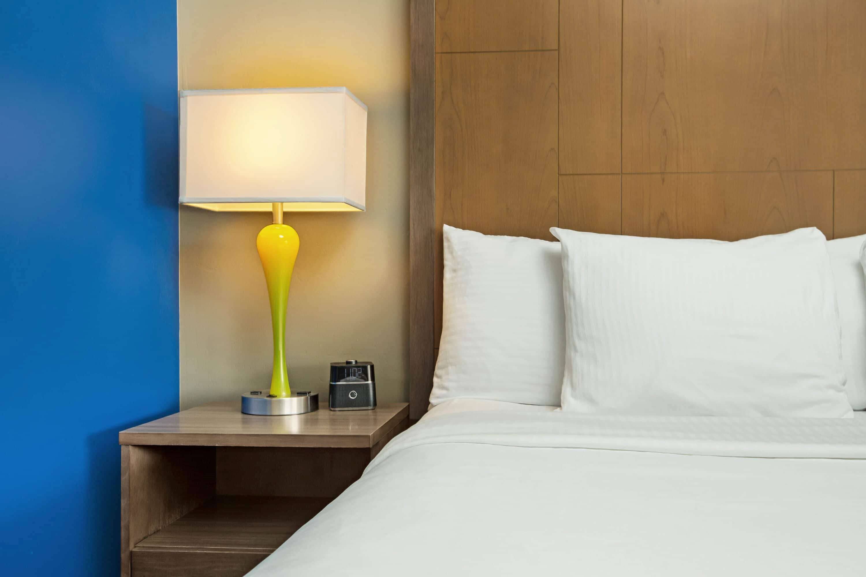 A hotel room with a bed, white pillows, a nightstand with a lamp, and a digital clock.