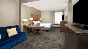 A hotel room with a blue sofa, a bed, a desk with lamps, a television, and a patterned carpet. - La Quinta Inn & Suites by Wyndham Page at Lake Powell (Page)