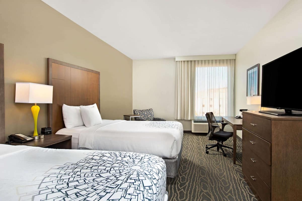 A hotel room with two beds, a desk, a chair, a television, and a lamp.