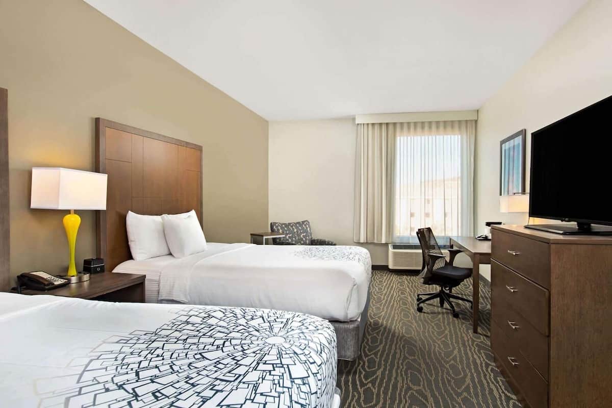 A hotel room with two beds, a desk, a chair, a television, and a lamp.