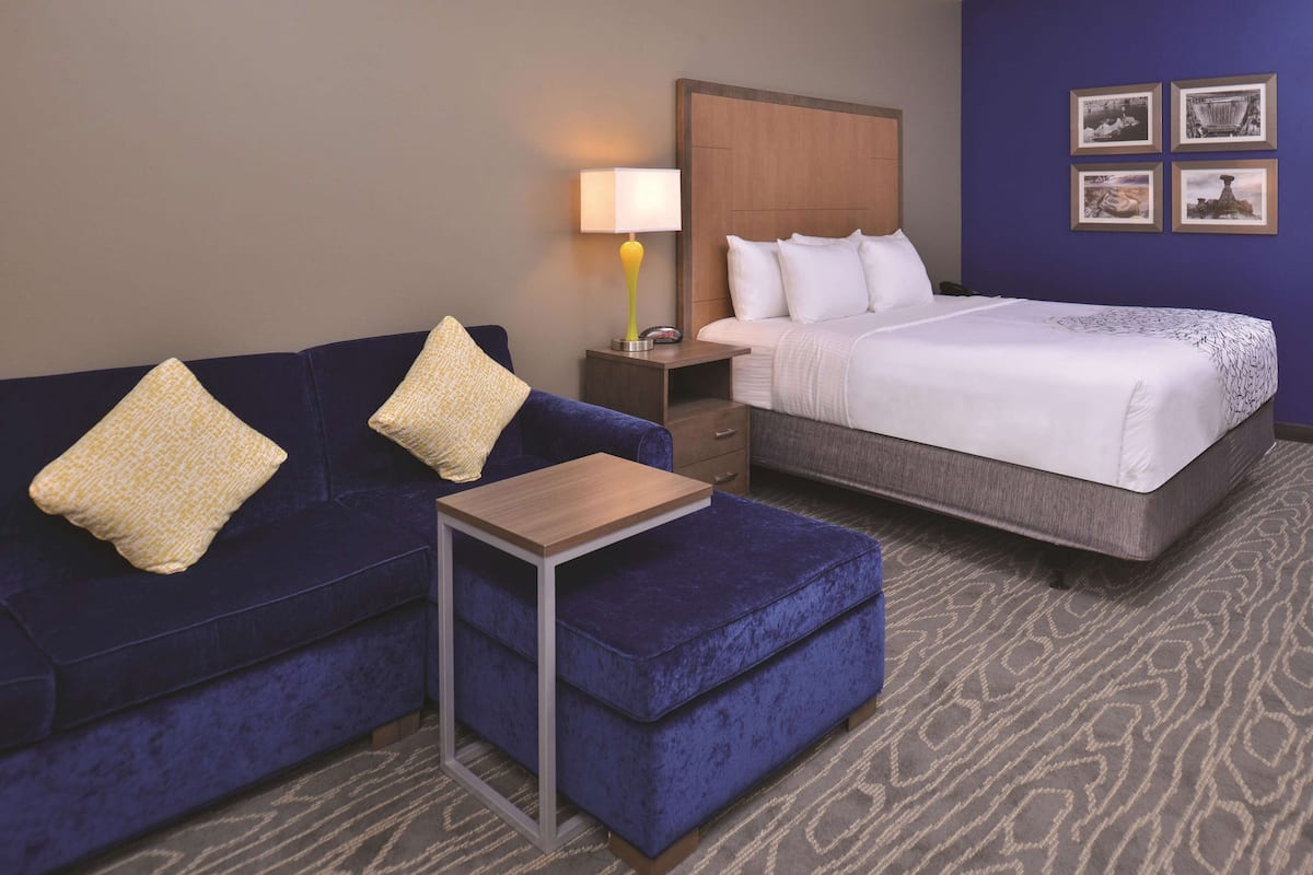 A hotel room with a blue sofa, a small table, a bed with white bedding, a nightstand with a lamp, and a wall with framed pictures.