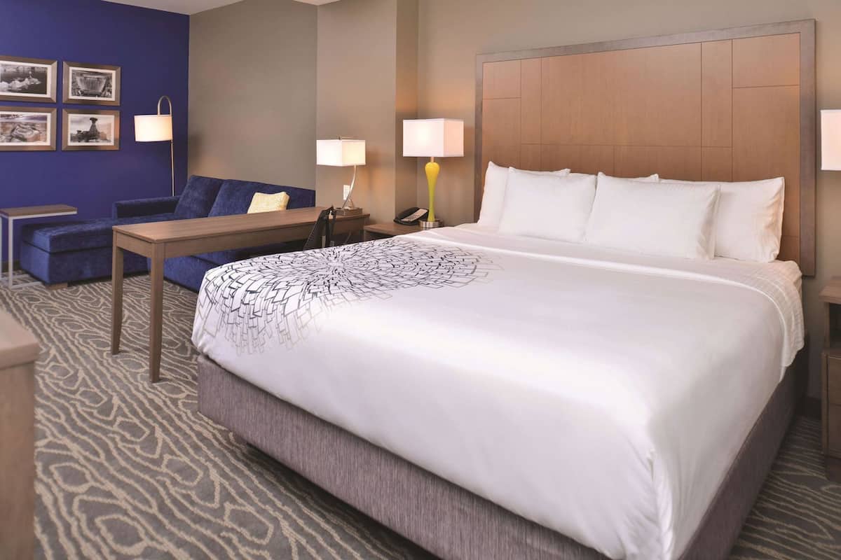 A hotel room with a large bed, a sofa, a small table, two lamps, and framed pictures on the wall.