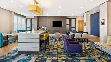 A modern hotel lobby with a bar area, seating, and a large flat-screen TV mounted on the wall.