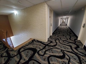 A hotel corridor with patterned carpet, a brick wall, and a wooden door. - Motel 6 Hastings, NE (Hastings)
