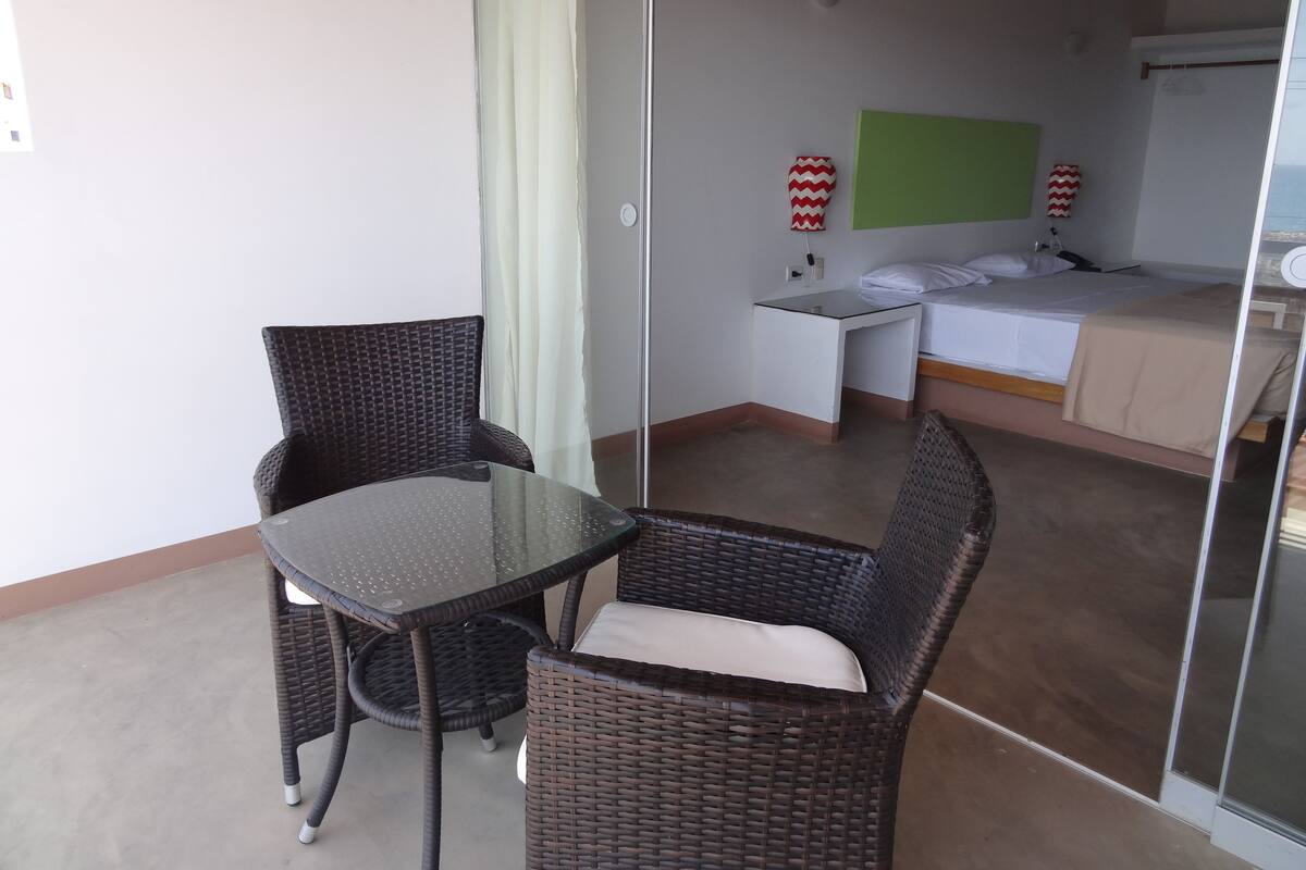A small, compact room with a glass partition separating the living area from the sleeping area. There is a single bed with white bedding, a small table with a chair, and two wall-mounted lamps with red and white zigzag patterns.