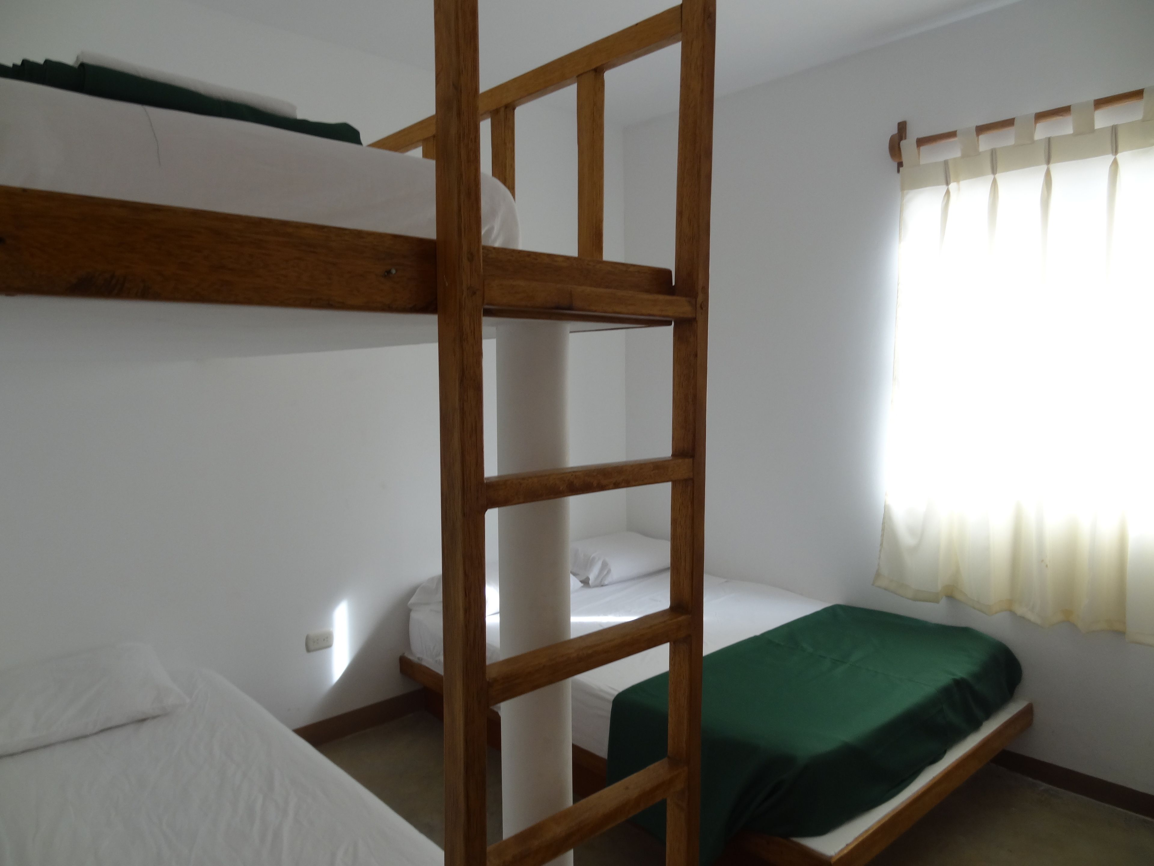A dormitory room with bunk beds, a window with curtains, and a wooden ladder.