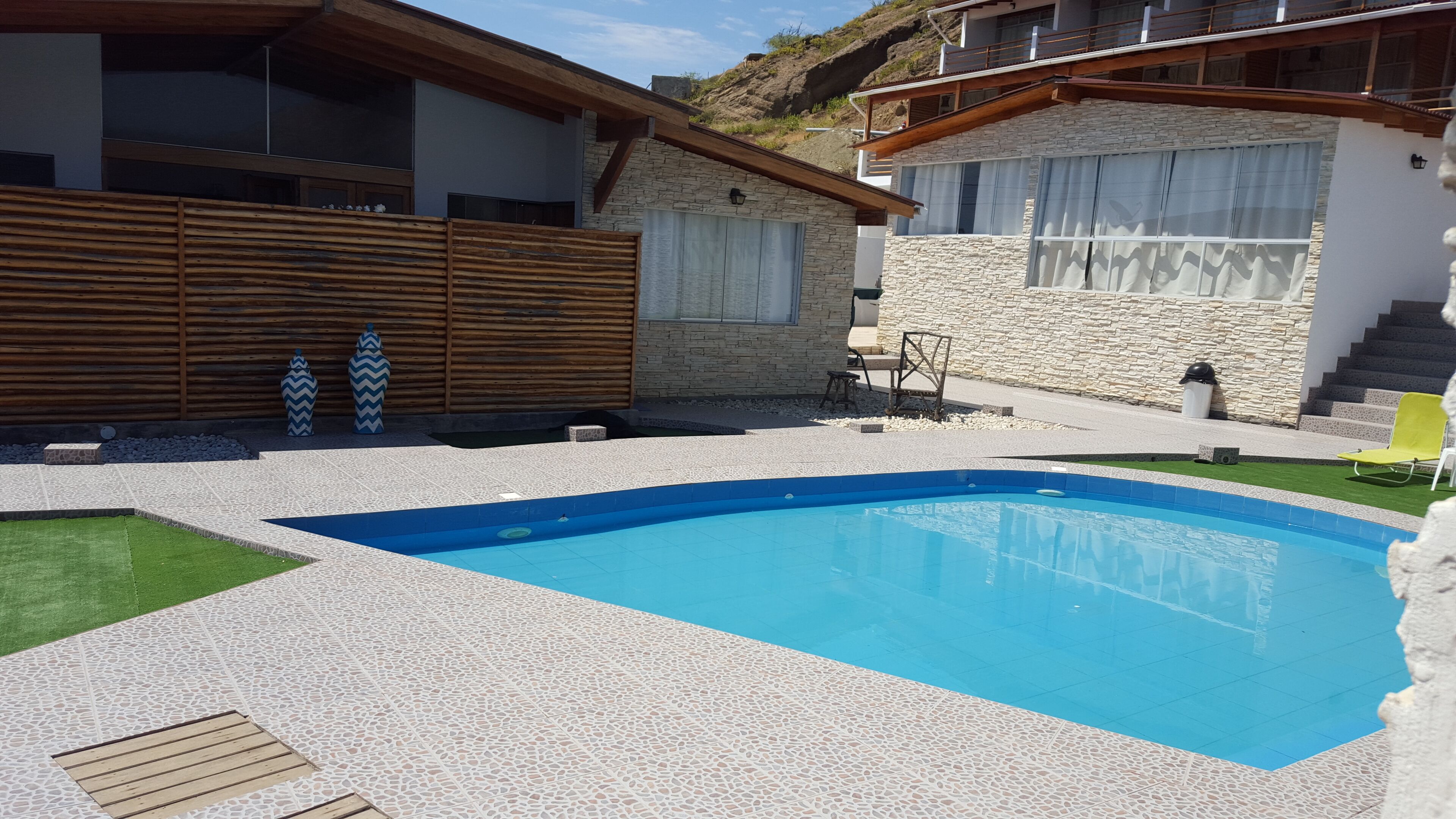 Un área de piscina moderna con muro de piedra, terraza de madera y una pequeña zona de estar al aire libre.