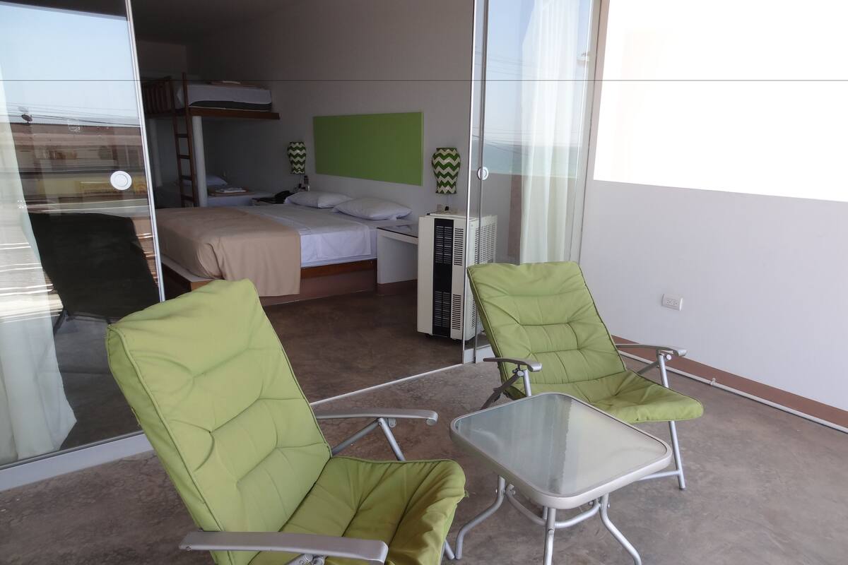 A hotel room with a bed, a desk, and two green armchairs with a glass table.