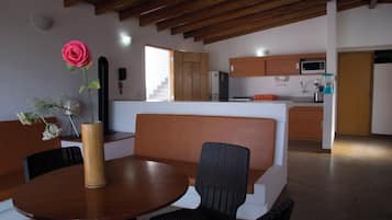 A dining area with a wooden table and chairs, a vase with a flower, and a kitchen in the background.