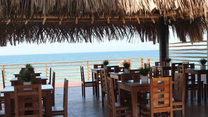 An outdoor dining area with wooden tables and chairs, potted plants, and a view of the ocean. - Akas Hotel Apartamentos (Canoas de Punta Sal)