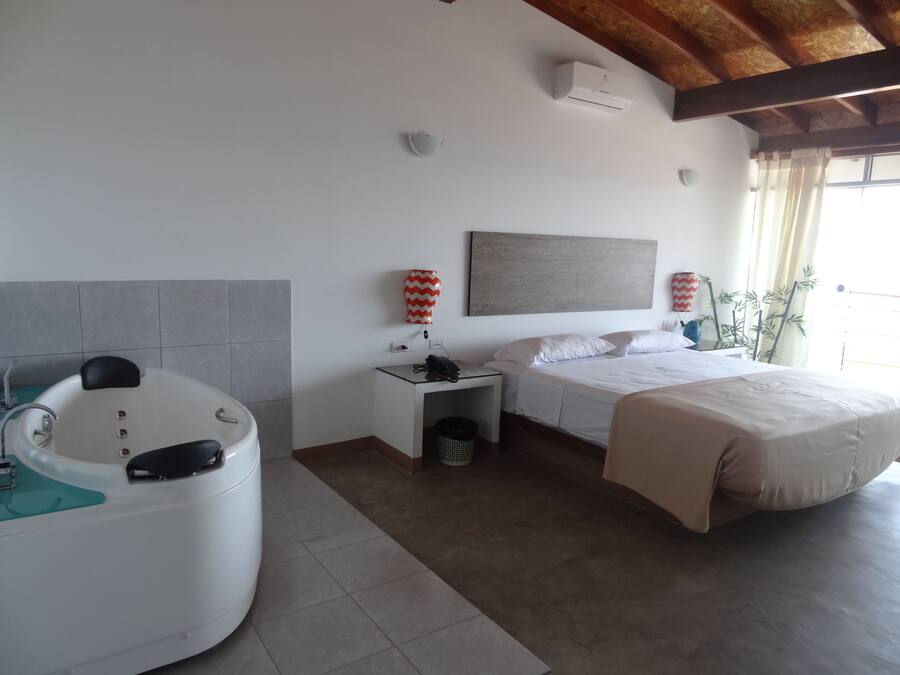 Habitación de hotel con bañera, cama, mesita de noche, calefactor en la pared y ventana con cortinas.