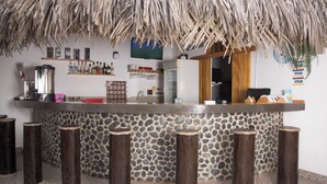 A bar area with a stone wall, wooden stools, a thatched roof, and a counter with various items. - Akas Hotel Apartamentos (Canoas de Punta Sal)