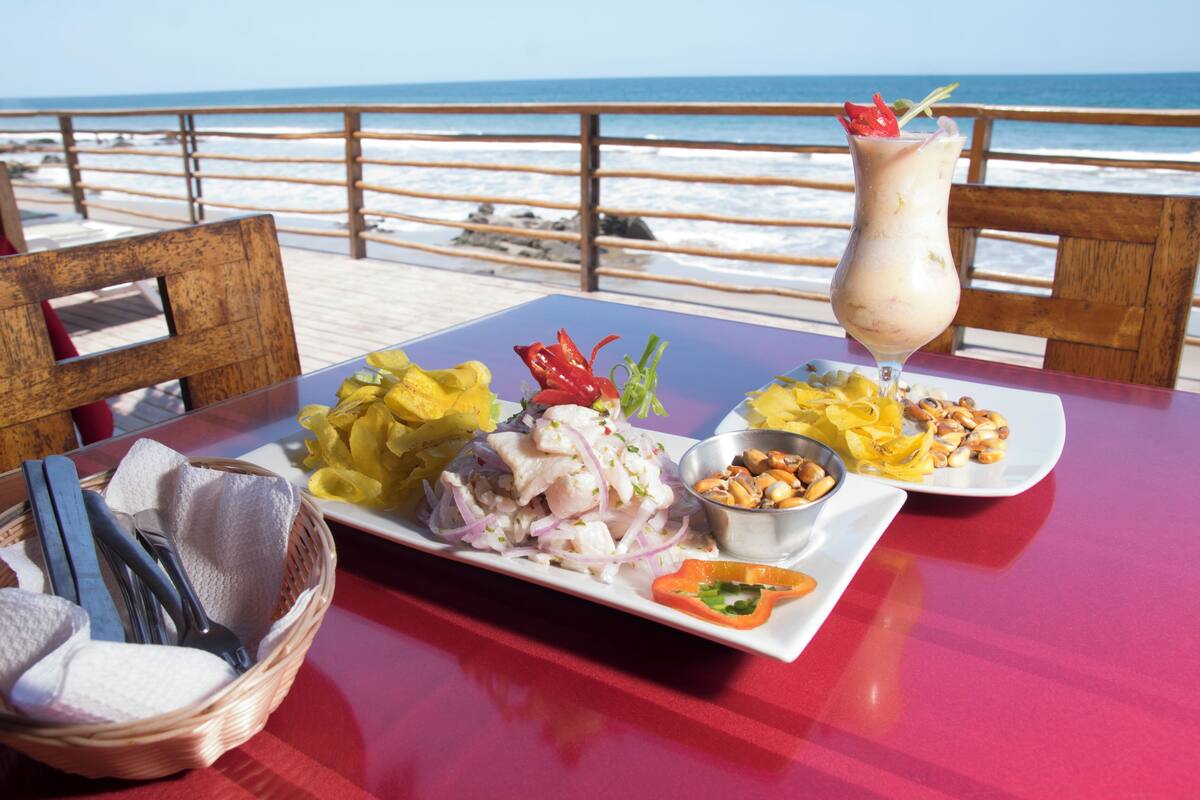 A table with a seafood dish, a drink, and a small bowl of nuts, set against a beach view.