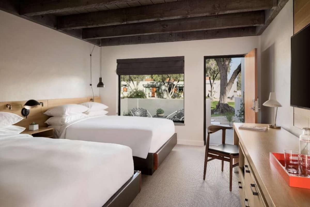 Une chambre d’hôtel avec deux lits, un bureau avec une chaise, une fenêtre donnant sur l’extérieur et une porte menant à un balcon.