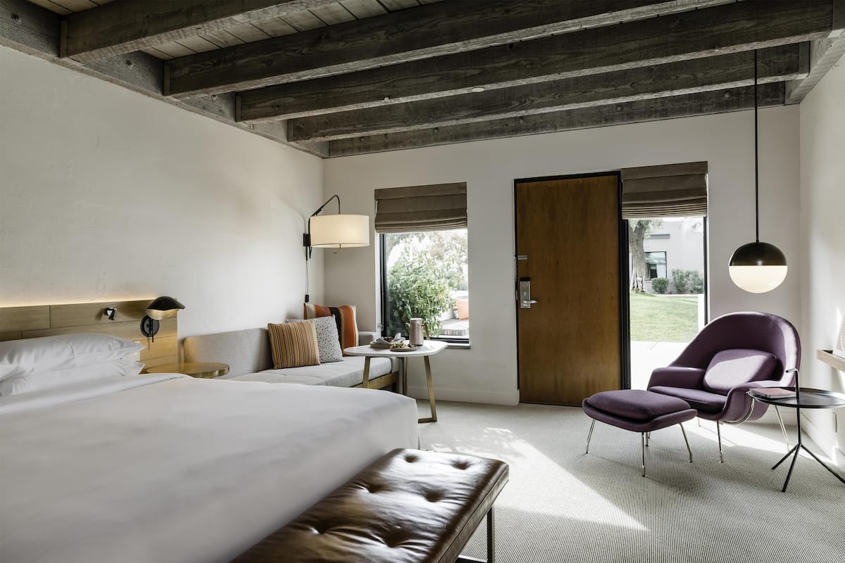 Une chambre d’hôtel moderne avec un grand lit, une petite table, une chaise et un banc. Il y a une fenêtre donnant sur l’extérieur.