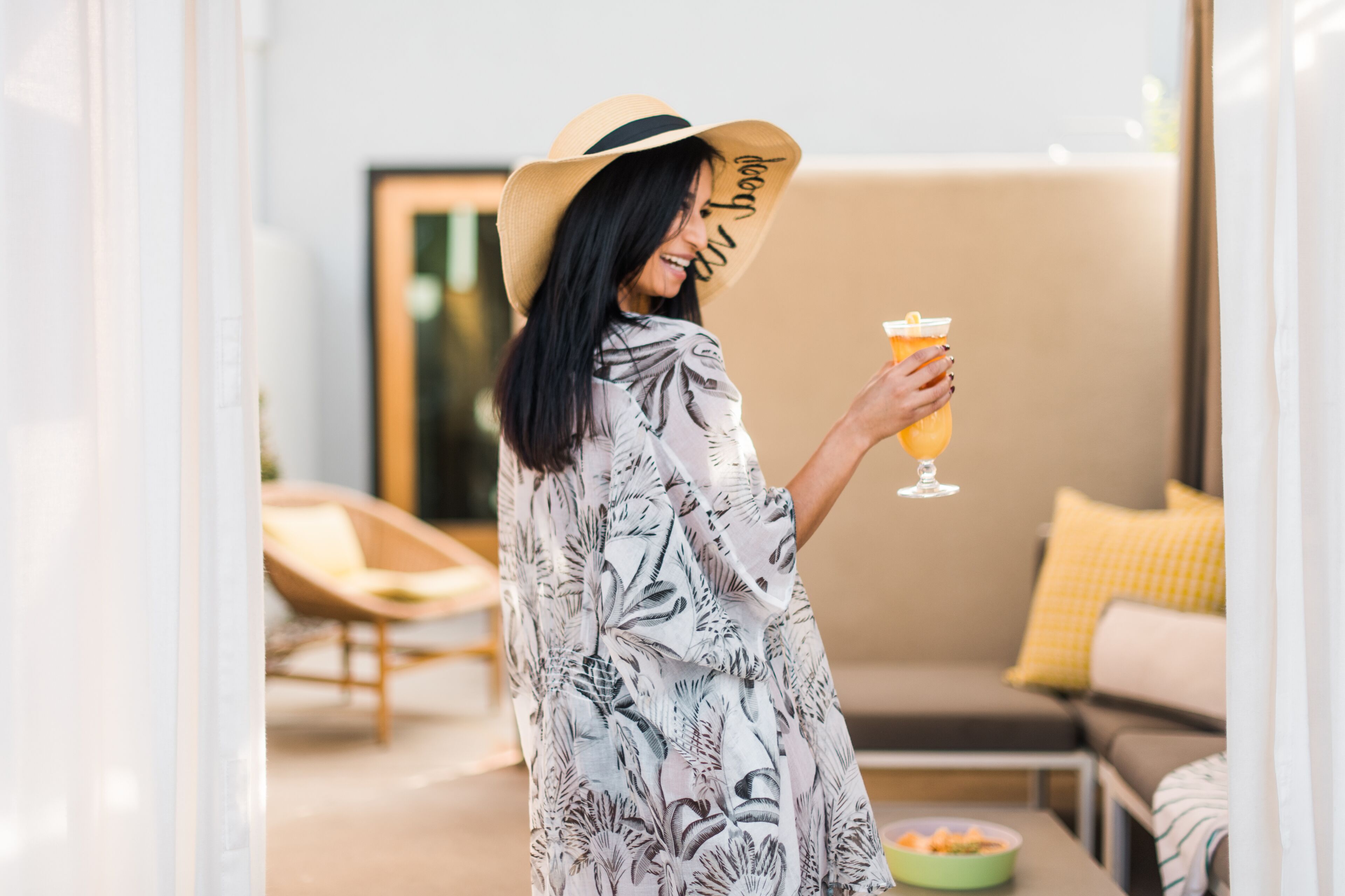A person in a patterned dress and hat holding a drink, standing in a modern living room with a sofa and a dining table.