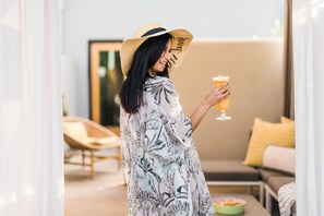 A person in a patterned dress and hat holding a drink, standing in a modern living room with a sofa and a dining table.