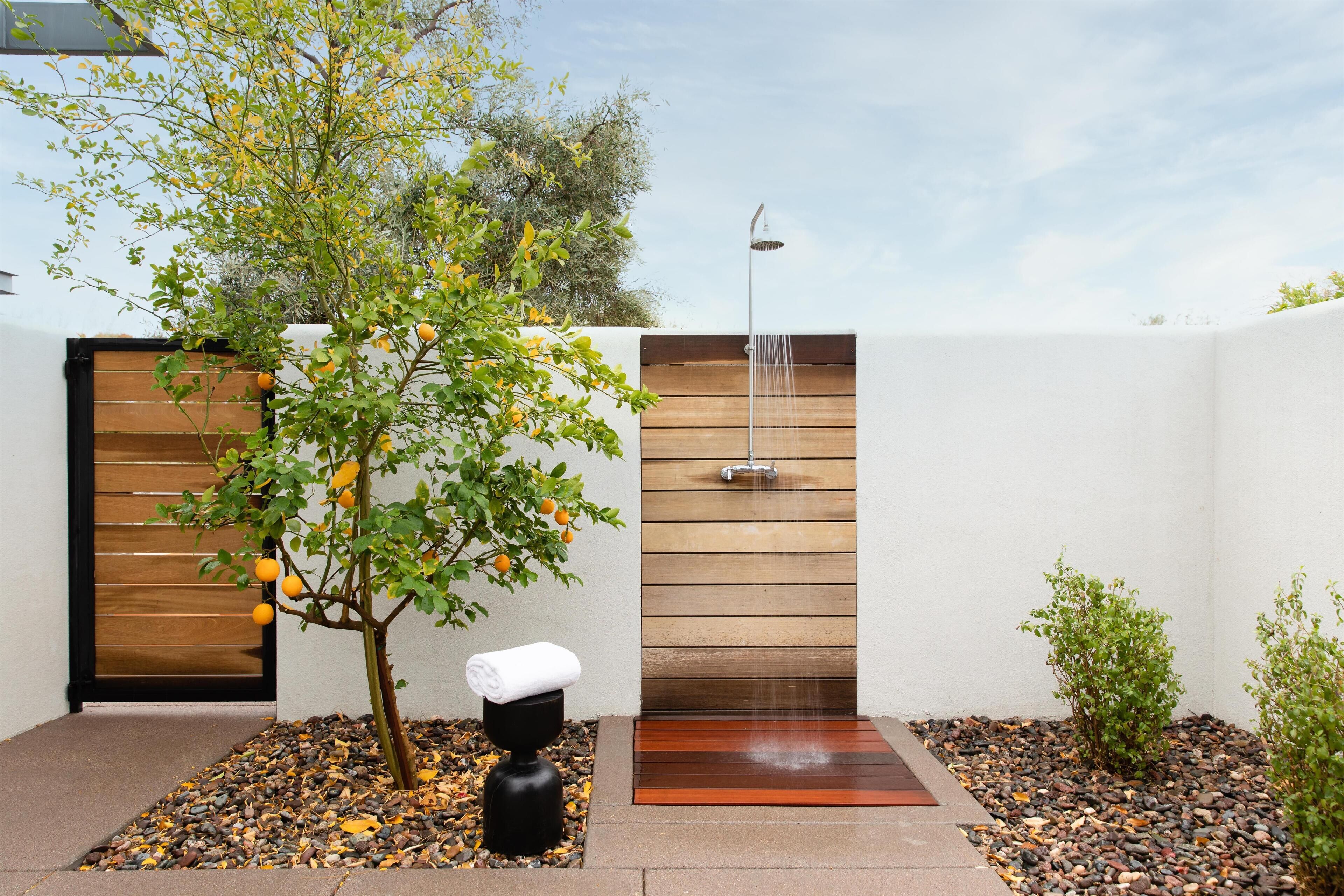 A modern outdoor shower area with a wooden wall, a tree with oranges, and a black sculpture.