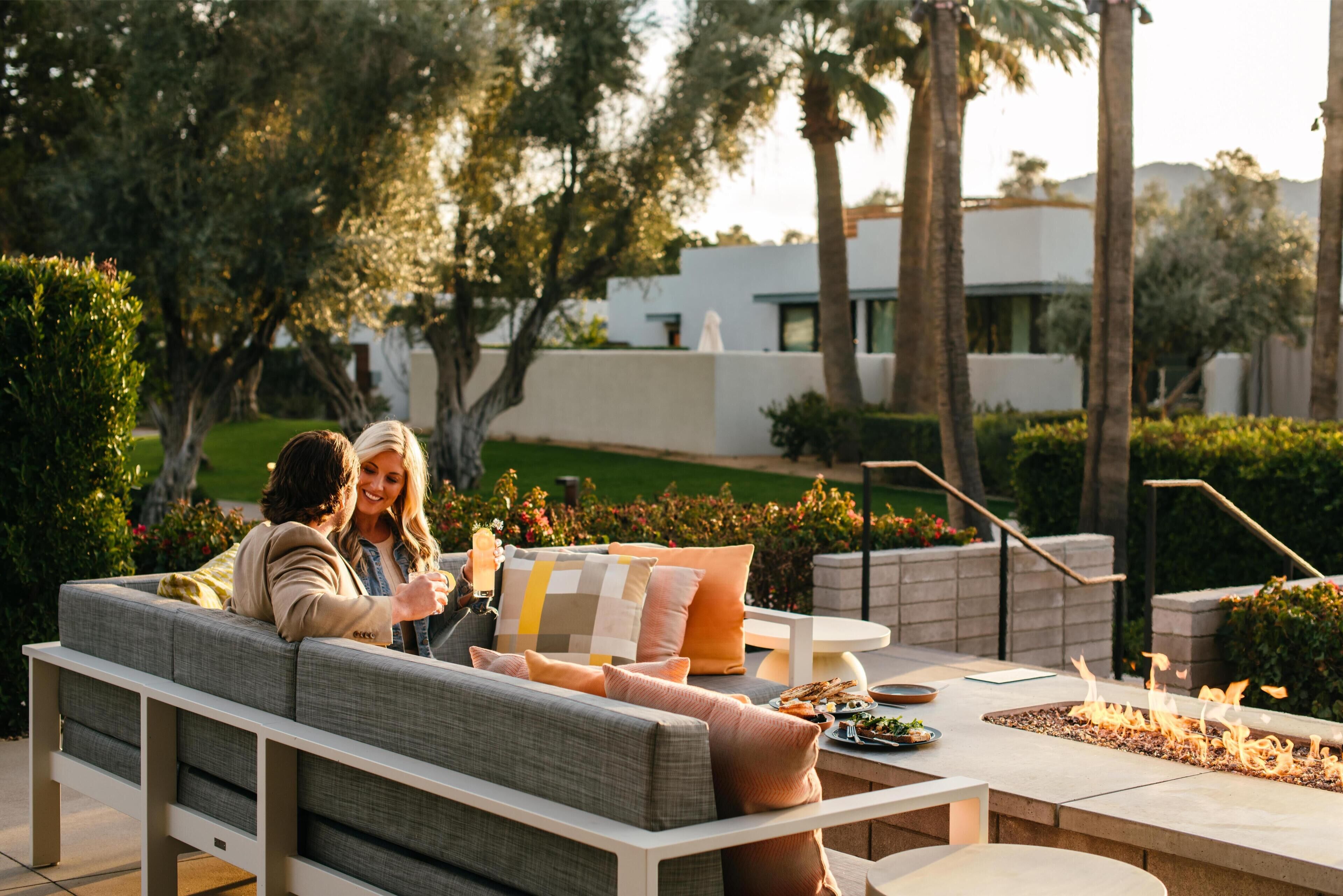 A modern outdoor seating area with a built-in fire feature, surrounded by greenery and a white building in the background.