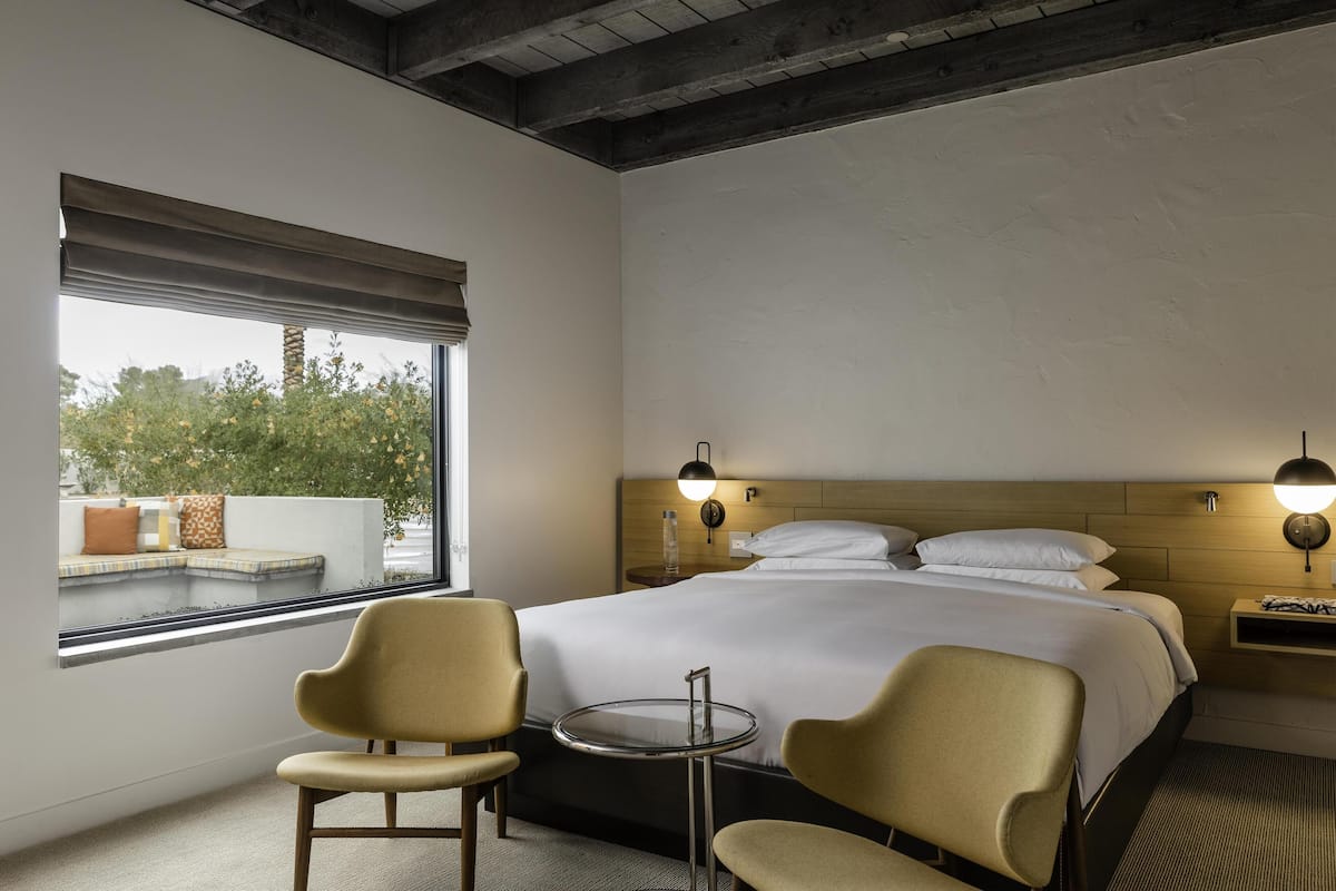 Une chambre d’hôtel avec un grand lit, deux tables de chevet, une petite table ronde, deux chaises et une fenêtre donnant sur un paysage verdoyant.