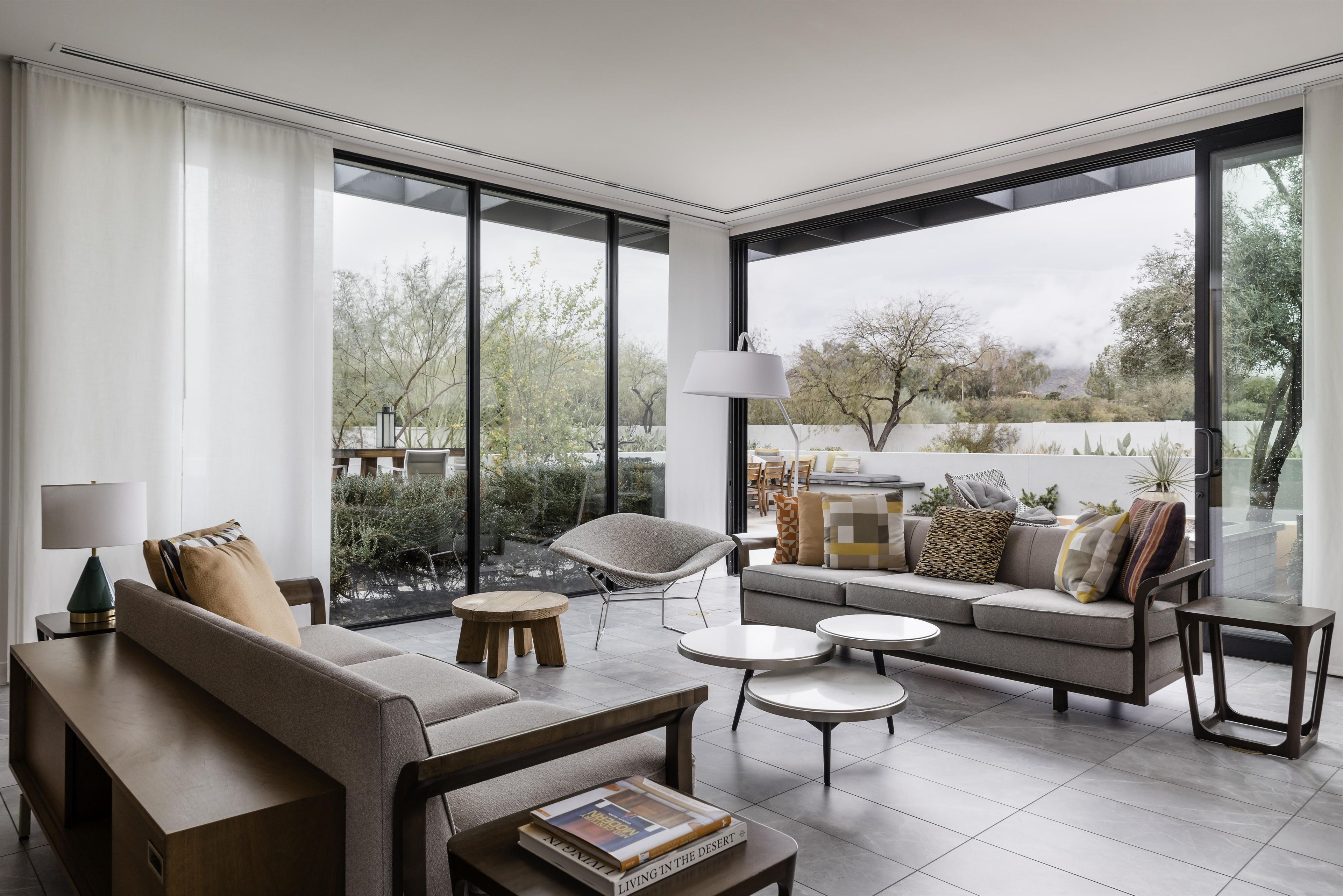 A modern living room with a sofa, coffee table, and chairs, large windows with a view of trees and a white fence.