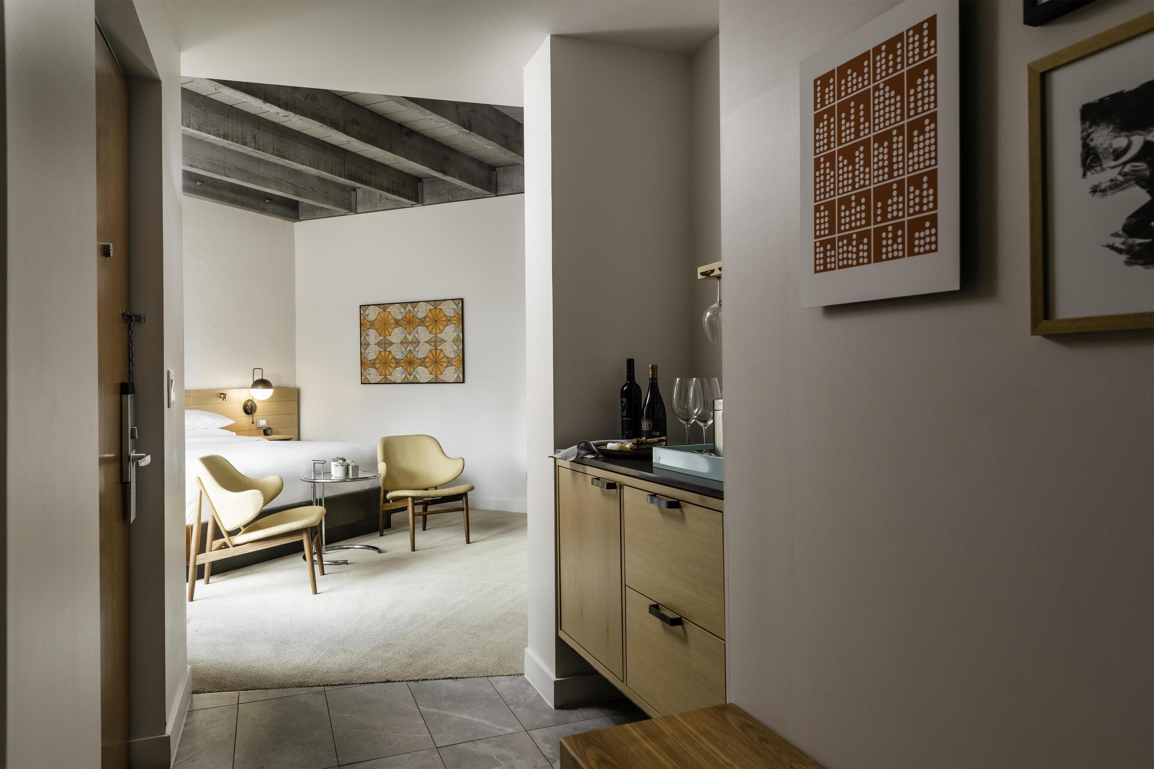 Una habitación de hotel con cama, una silla, escritorio, armario y obras de arte en las paredes.