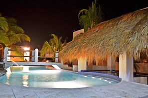 A swimming pool with a thatched roof structure, palm trees, and a night-time setting. - Playa Plana Hotel (Chiquimulilla)