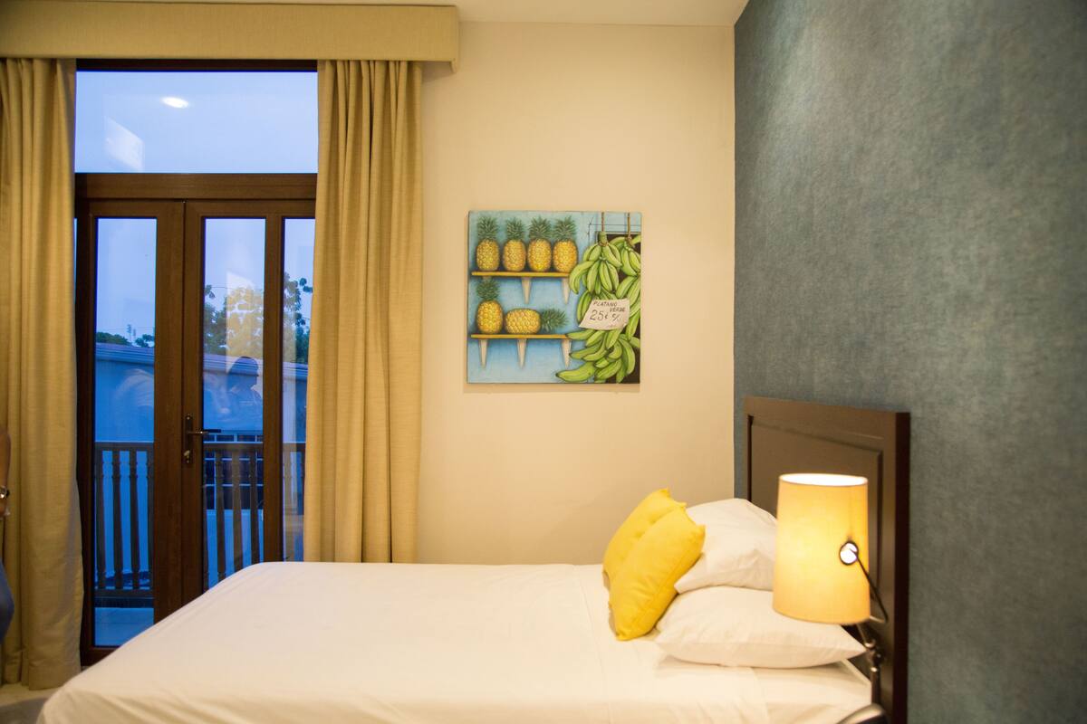 Una habitación de hotel con una cama, una mesita de noche con lámpara, una ventana con cortinas y una obra de arte con piñas y plátanos.