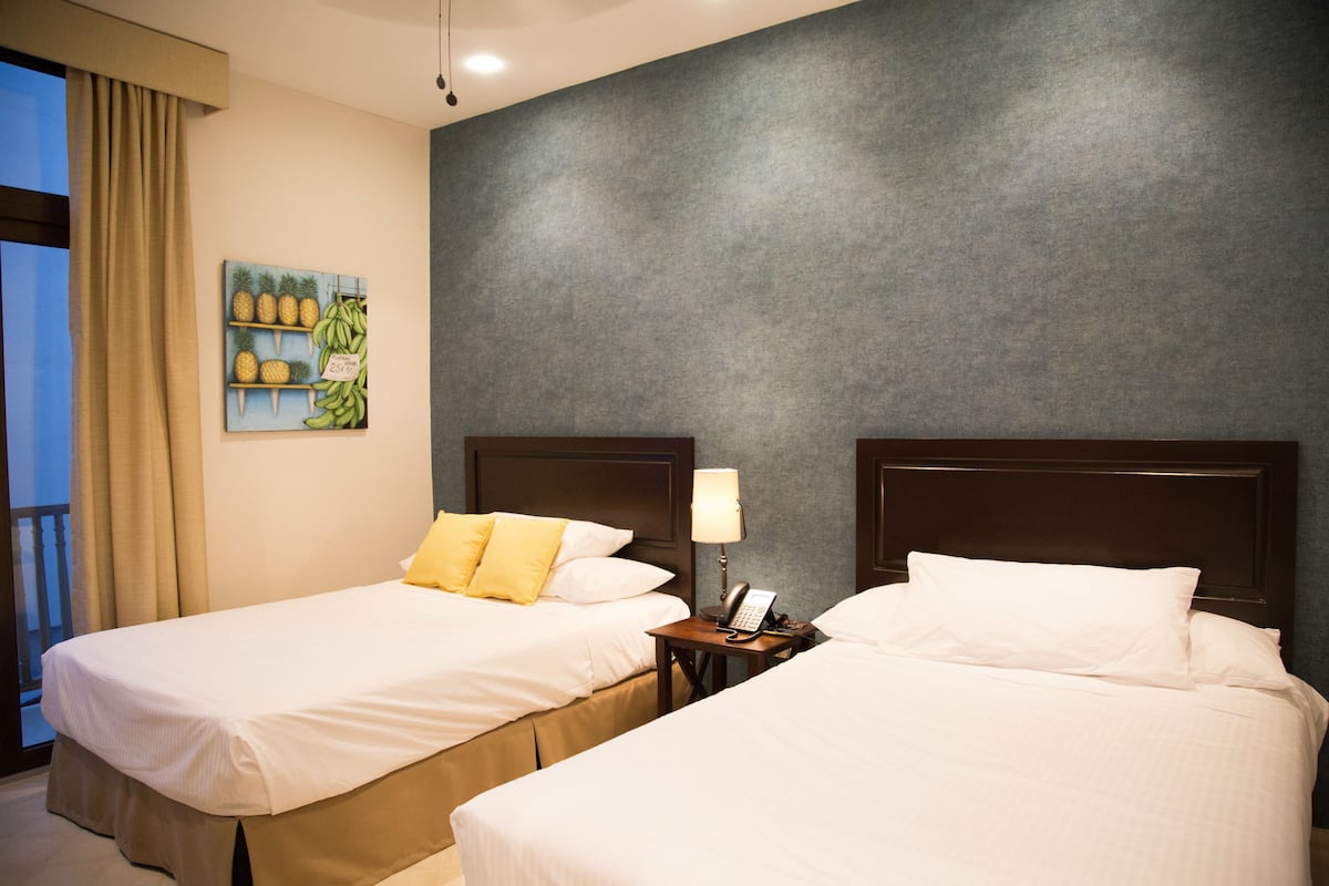 Habitación de hotel con dos camas, una mesita de noche con lámpara y un cuadro de frutas en la pared.